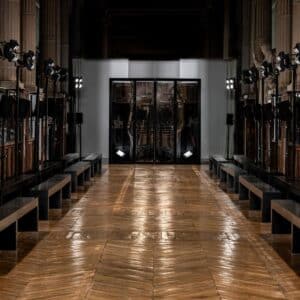 Backstage area of a theater or studio with black benches along narrow wooden floor, walls lined with lights and equipment, and glass doors at the far end.